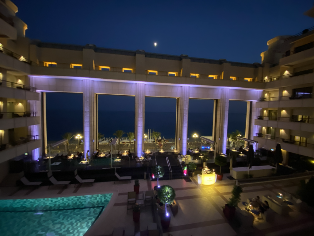 Hotel courtyard at dusk with a pool and outdoor dining, looking out through large arches to the Mediterranean
